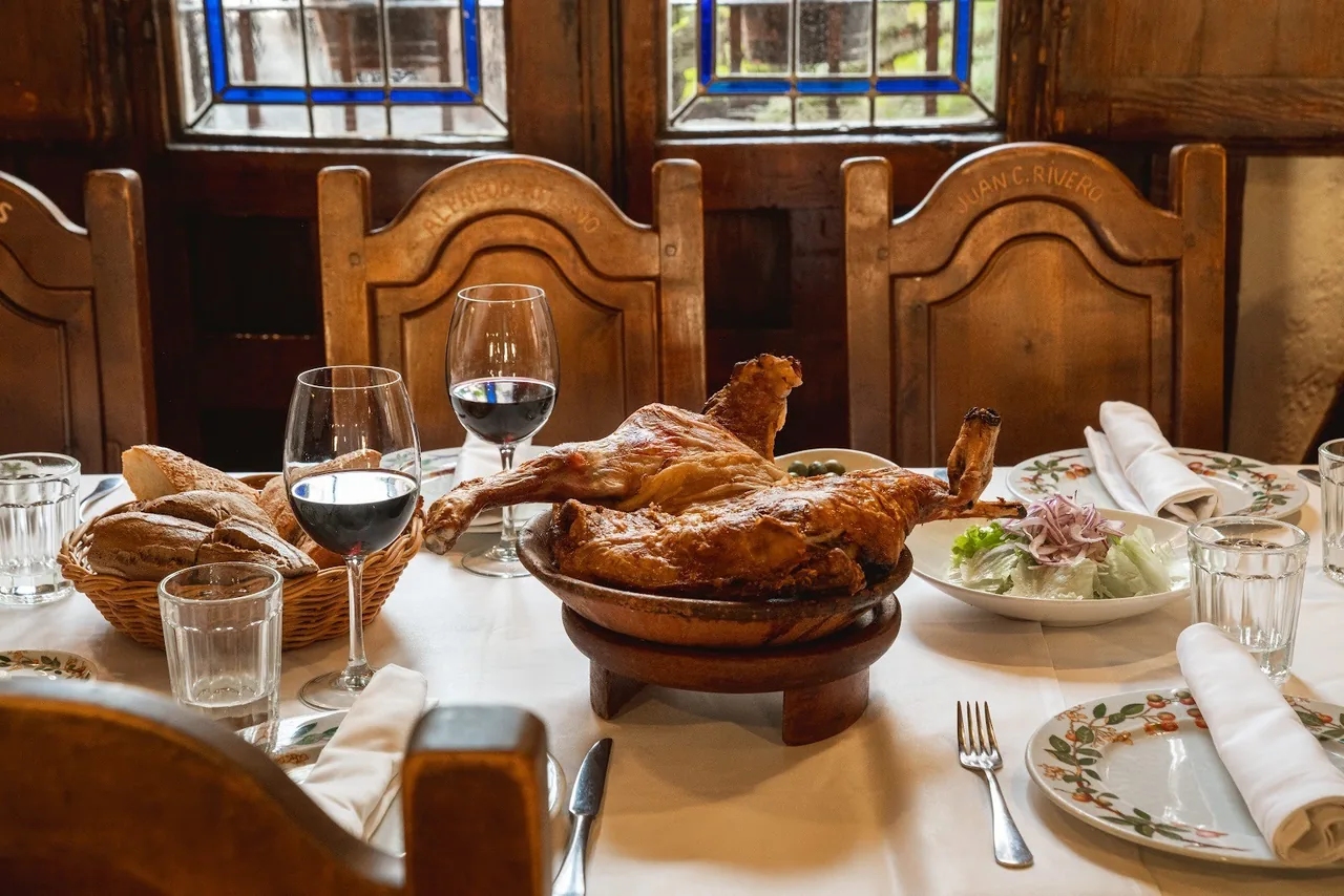A table set with a roast lamb in a clay dish, a glass of red wine and a side salad, beneath a stained-glass window