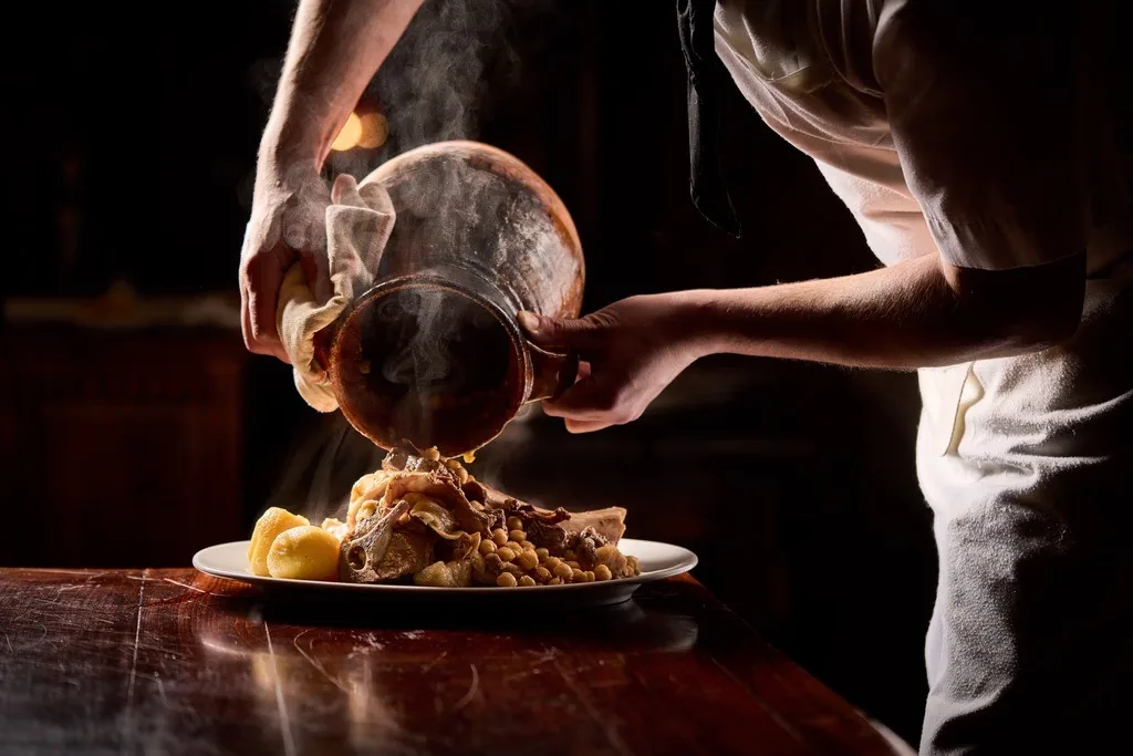 A cook lifting a clay pot over a plate of boiled meats, potatoes and chickpeas as steam rises