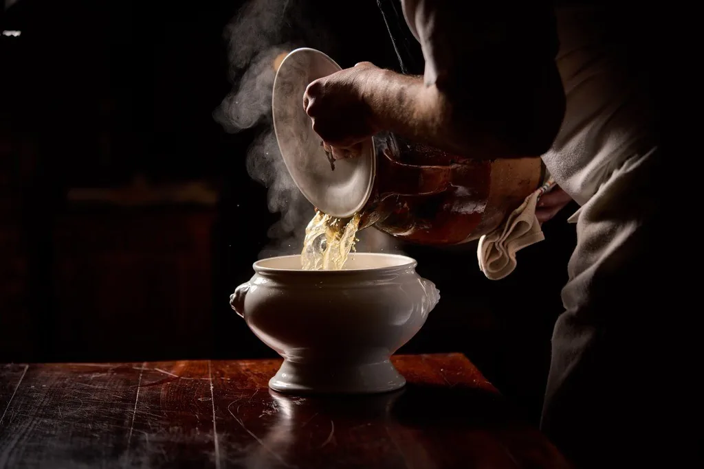 Steam rising as a cook pours noodle broth into a white tureen on a dark table