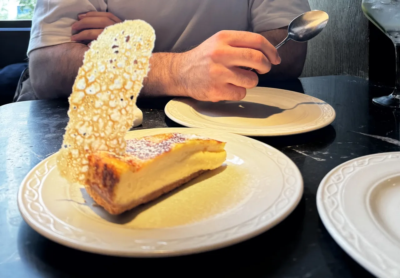 A slice of baked cheesecake on a cream plate with a thin lace-like sugar tuile standing upright beside it