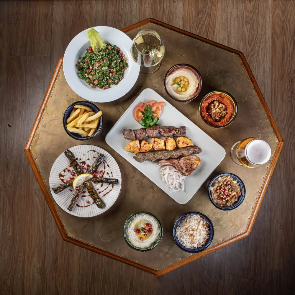 Overhead Lebanese-style mezze spread on a wood-and-brass table: tabbouleh, hummus, muhammara, stuffed vine leaves, mixed grill skewers, fries, plus a glass of wine and a beer