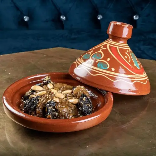 Moroccan lamb tagine with prunes, sesame and pine nuts in a terracotta dish, its painted conical lid set beside it against a blue velvet backdrop