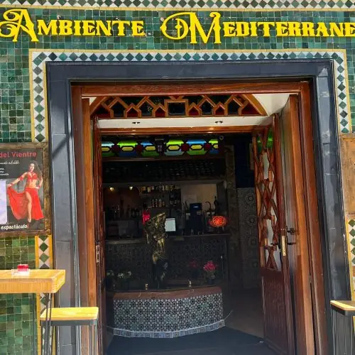 Restaurant entrance with an ornate green-and-gold Moroccan tiled facade reading 'Ambiente Mediterraneo', wooden doors open to a tiled bar counter and stained glass inside