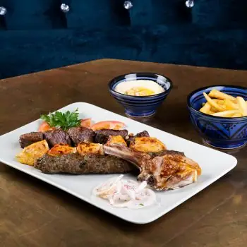 Mixed grill platter with kofta, chicken and a lamb chop, tomato and onion garnish, plus a bowl of fries and a bowl of dip on a brass table