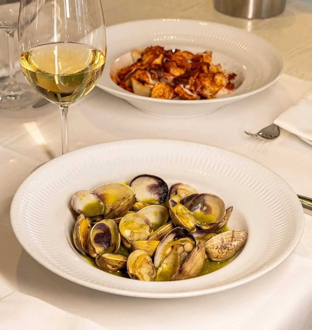 A bowl of clams in broth and a plate of octopus beside a glass of white wine