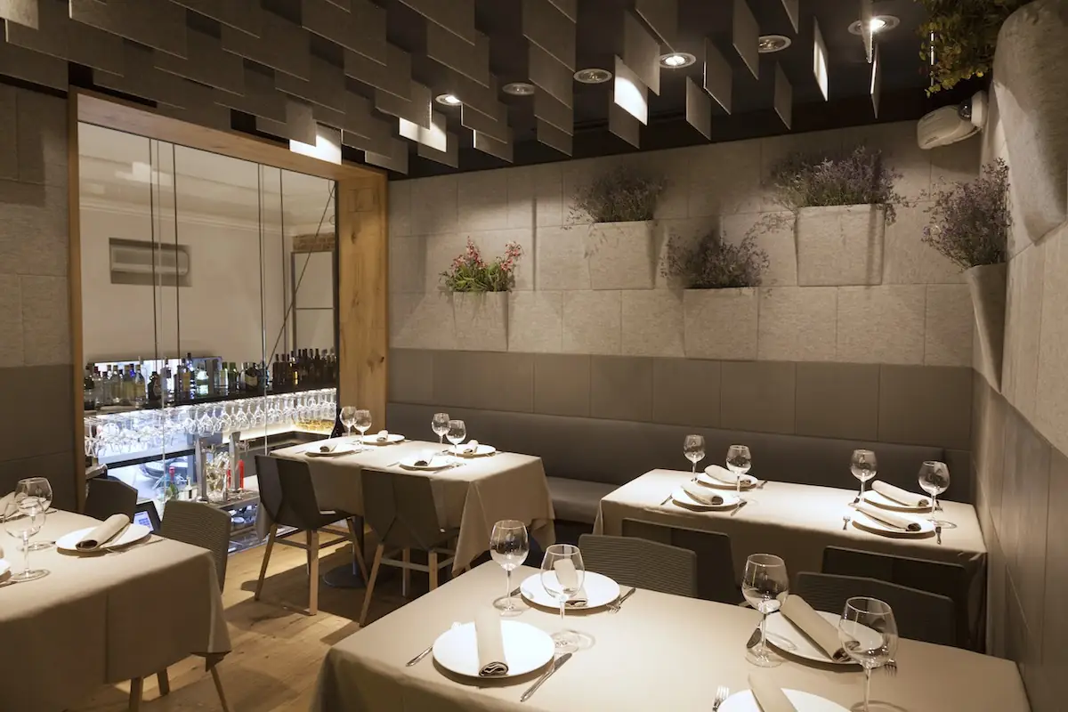 Dining room with tables set in cream linen and wine glasses, banquette seating, wall planters of dried flowers and a mirrored bar