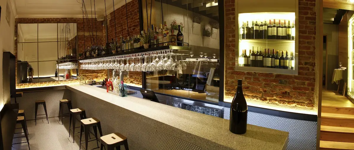 Long bar with exposed brick wall, backlit wine-bottle niches, rows of hanging stemware and wooden stools along a terrazzo counter