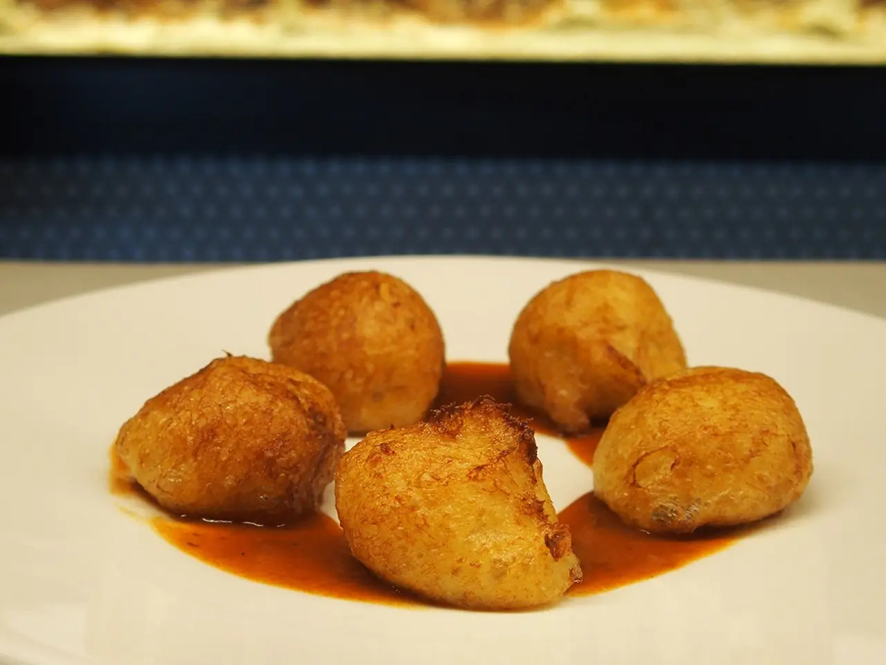 Five golden fried croquettes or fritters sitting in a pool of orange-red sauce on a white plate
