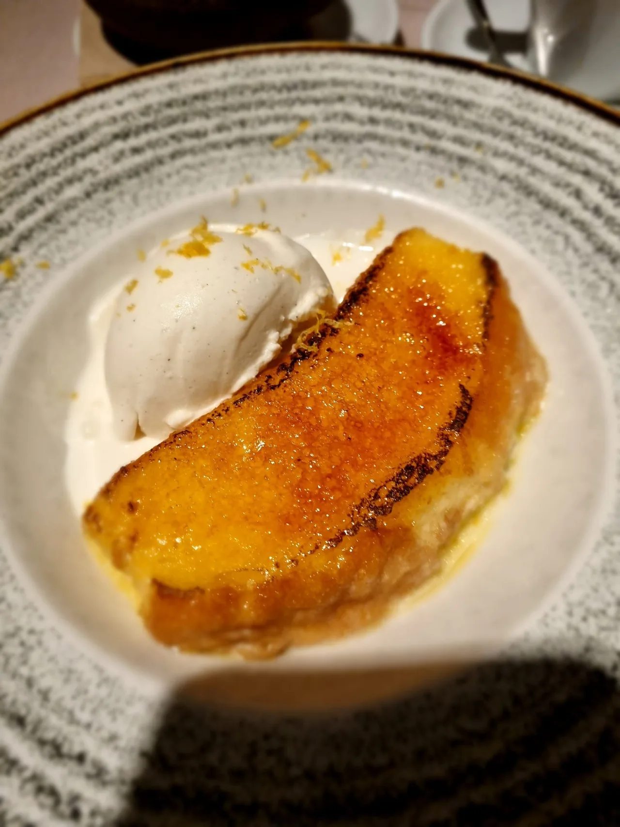 A caramelised golden slice of toast beside a scoop of ice cream with lemon zest on a grey speckled plate