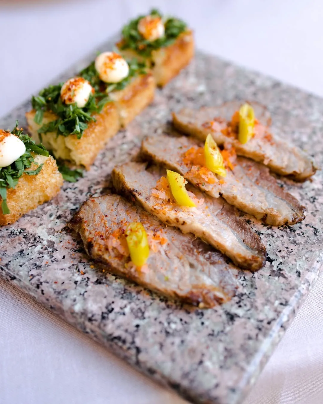 Sliced roast meat with small toppings on a stone-effect board next to toasts