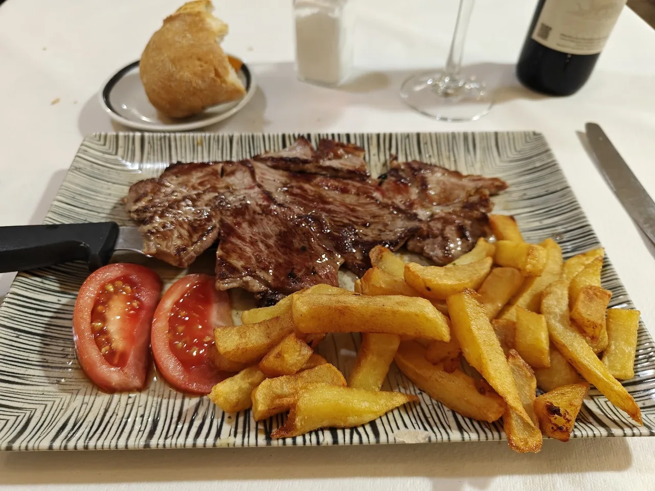 A striped plate of sliced grilled steak with fried potatoes and two tomato slices