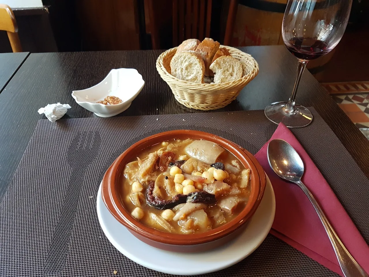 A terracotta bowl of stew with white beans, with a basket of bread and a glass of wine
