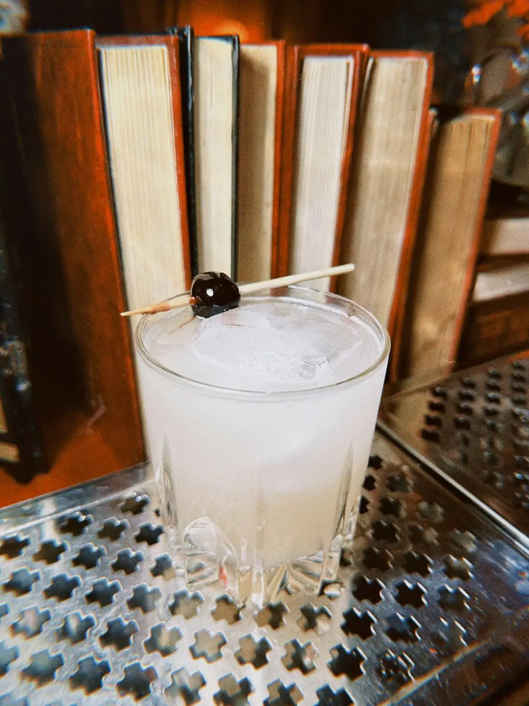 Clear cocktail on ice with a cherry garnish, served on a metal bar top