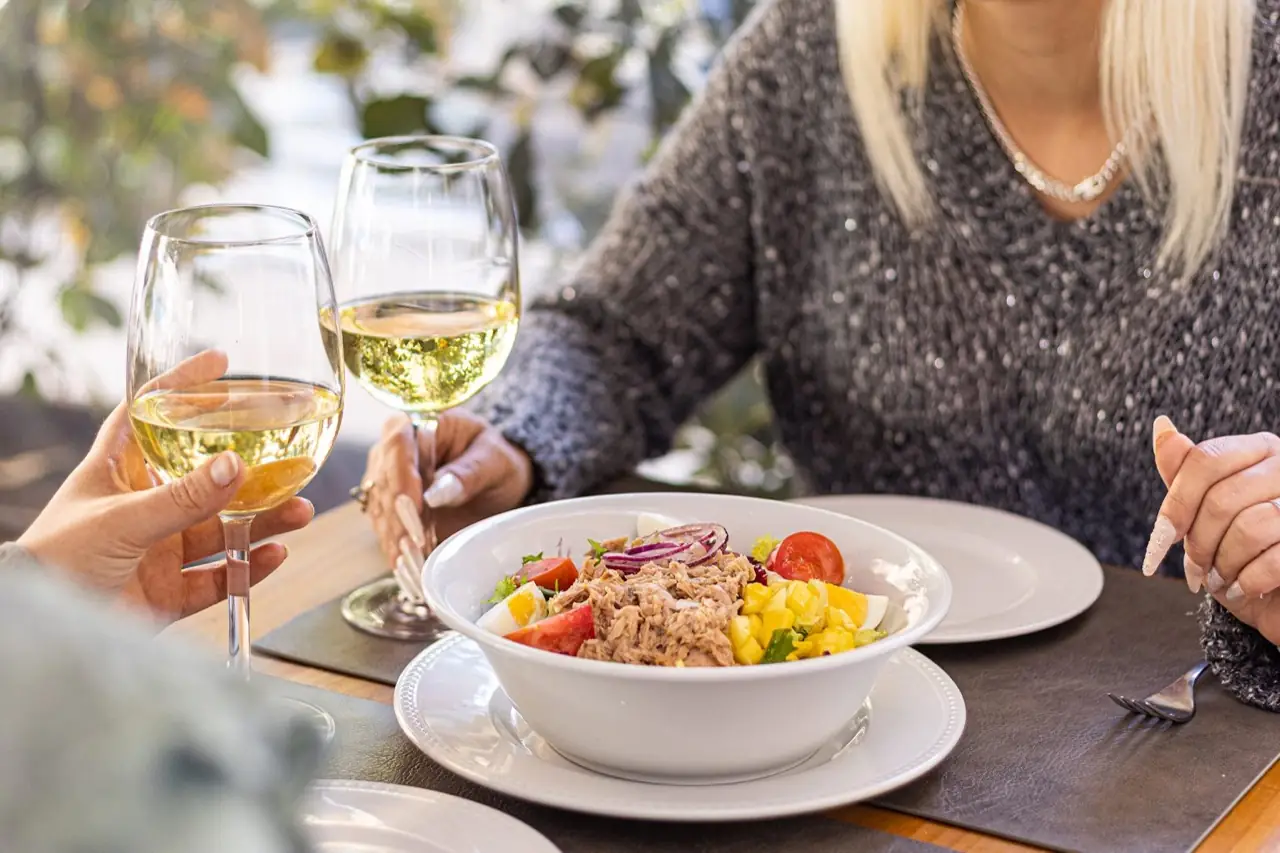 Two diners toasting glasses of white wine over a bowl of tuna salad with tomato, egg and mango on a set table