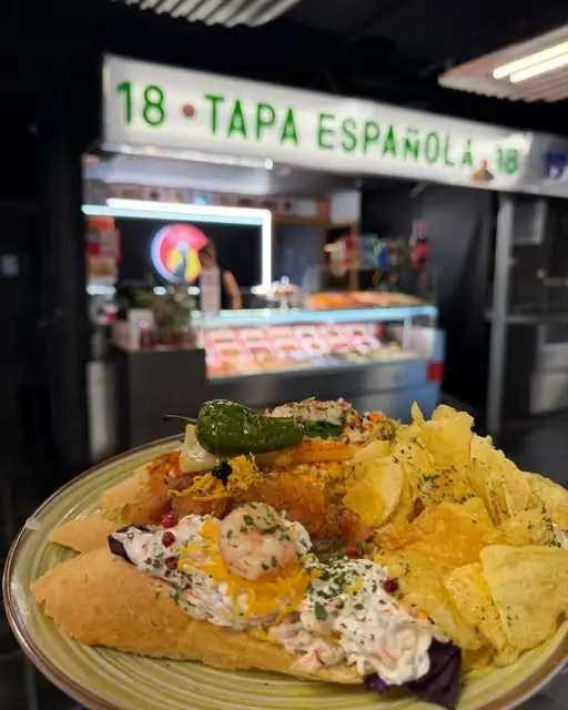 Open sandwich piled with shrimp salad, salmon, peppers and potato chips on a green plate, with the market stall sign reading 'Tapa Espanola' behind