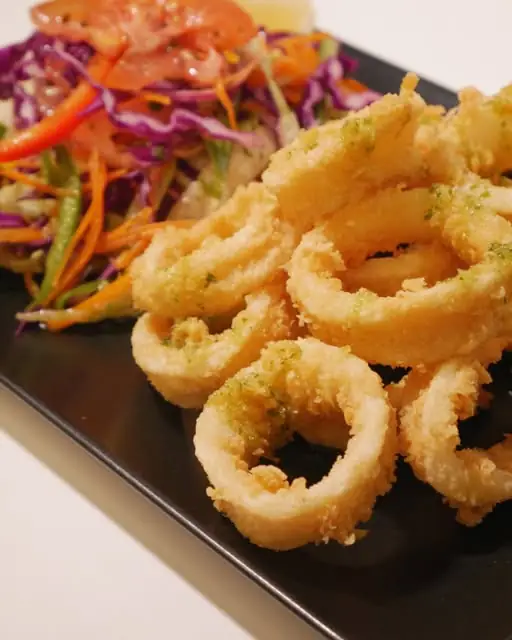 Battered fried calamari rings dusted with green herbs beside a shredded vegetable salad on a black rectangular plate