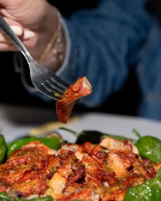 Fork lifting a piece of grilled octopus over a plate of octopus with padron peppers and green sauce