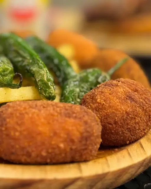 Two breaded croquettes with fried padron peppers and fries on a wooden plate