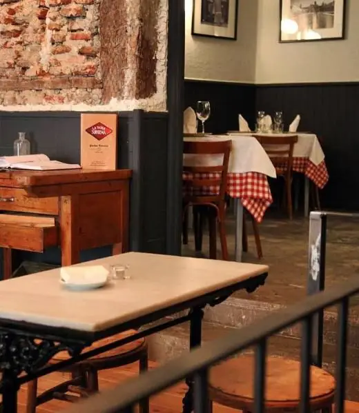 Dining room with exposed brick wall, dark wainscoting, and bistro tables dressed in red-and-white gingham cloths and bentwood chairs