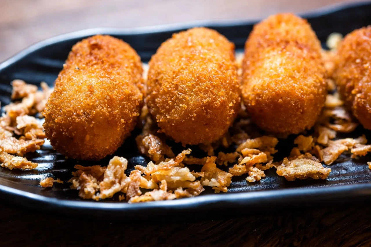 Four golden croquetas on a dark tray with a crumb garnish