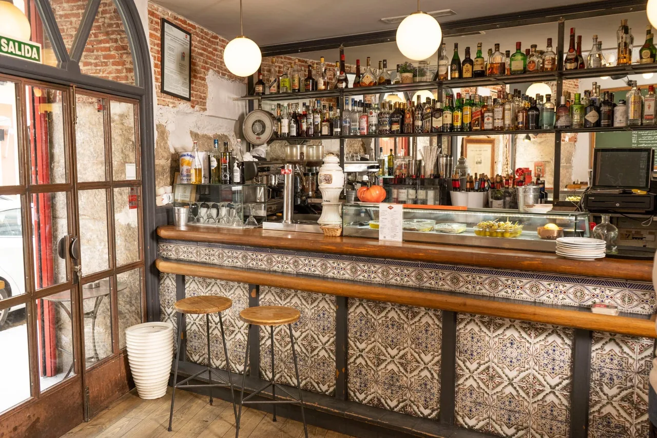 A tiled bar counter with a wooden top, brick walls, stools and shelves of bottles behind.