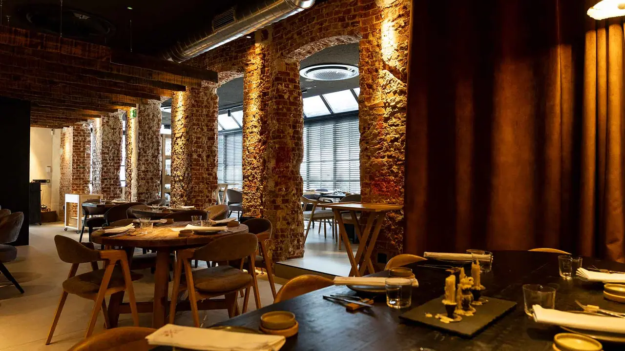 Dining room with exposed rubble-and-brick columns lit from below, a heavy rust-coloured curtain to one side, round and rectangular wooden tables laid with napkins and glasses, and daylight coming through slatted blinds at the back