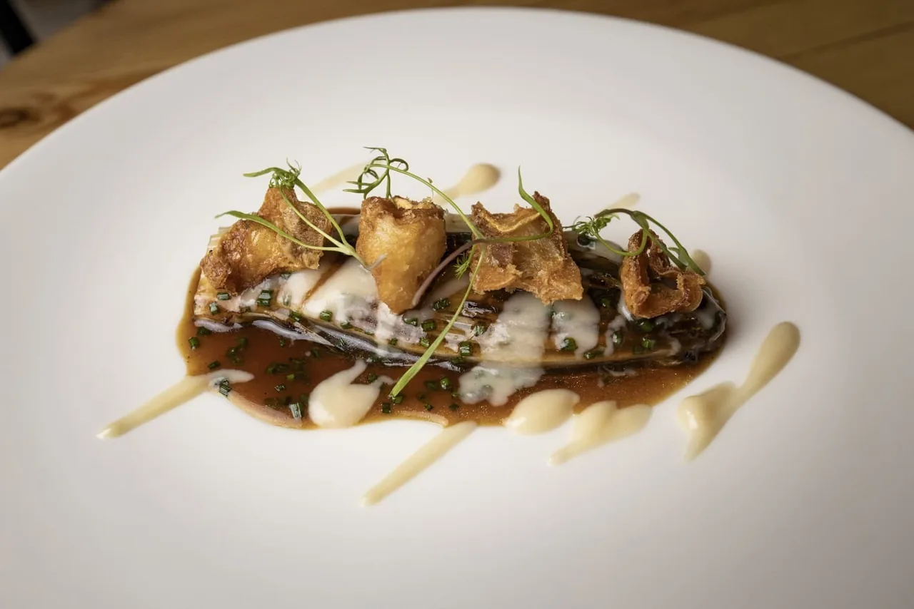 A piece of fish topped with crisp mushrooms and herbs on a dark sauce on a white plate