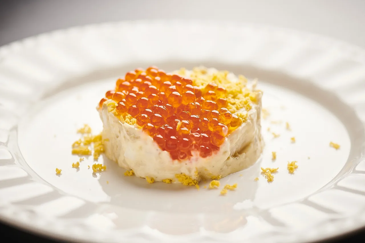 A round dish topped with bright orange roe and yellow crumbs on a white plate