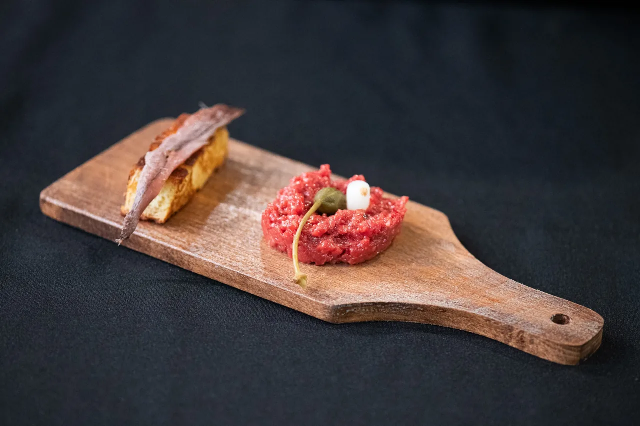 A wooden board with an anchovy on toast and a small mound of red tartare on a dark cloth