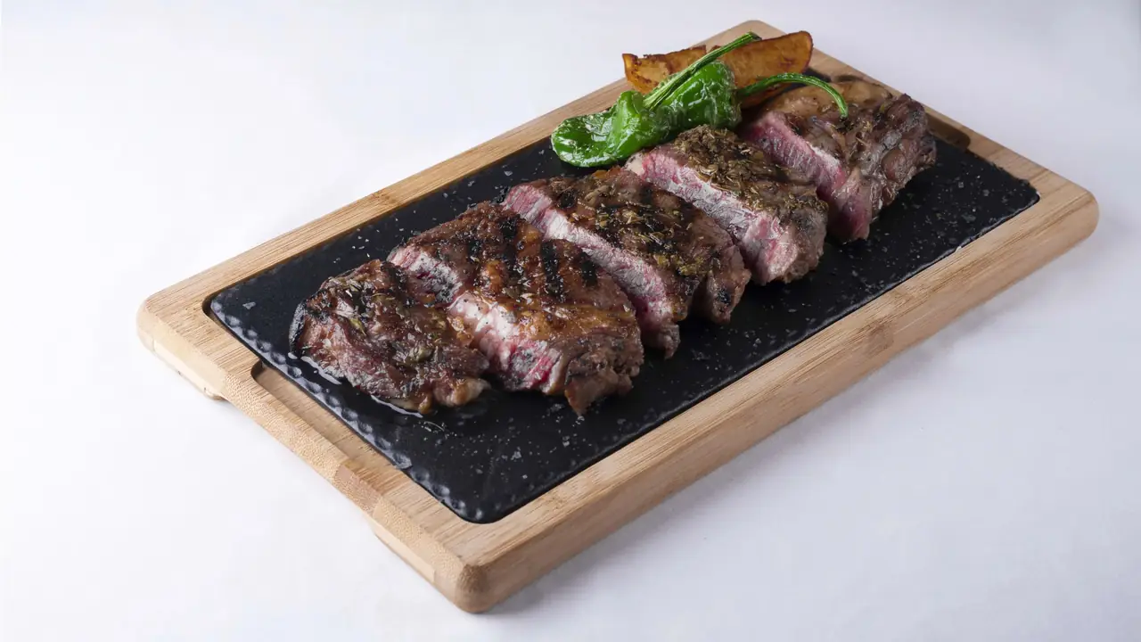 Grilled sliced steak with charred green padrón peppers and roasted potato wedges on a black slate board, La Tata