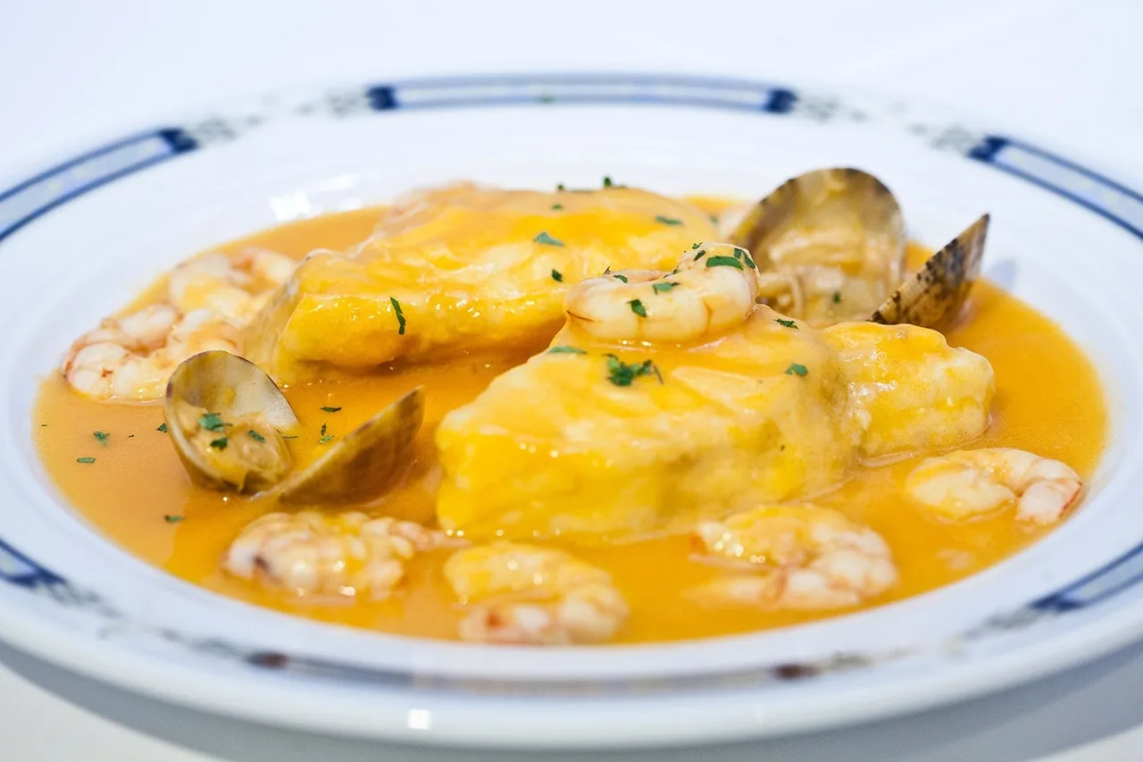 A piece of fish in orange sauce with clams and prawns in a blue-rimmed dish