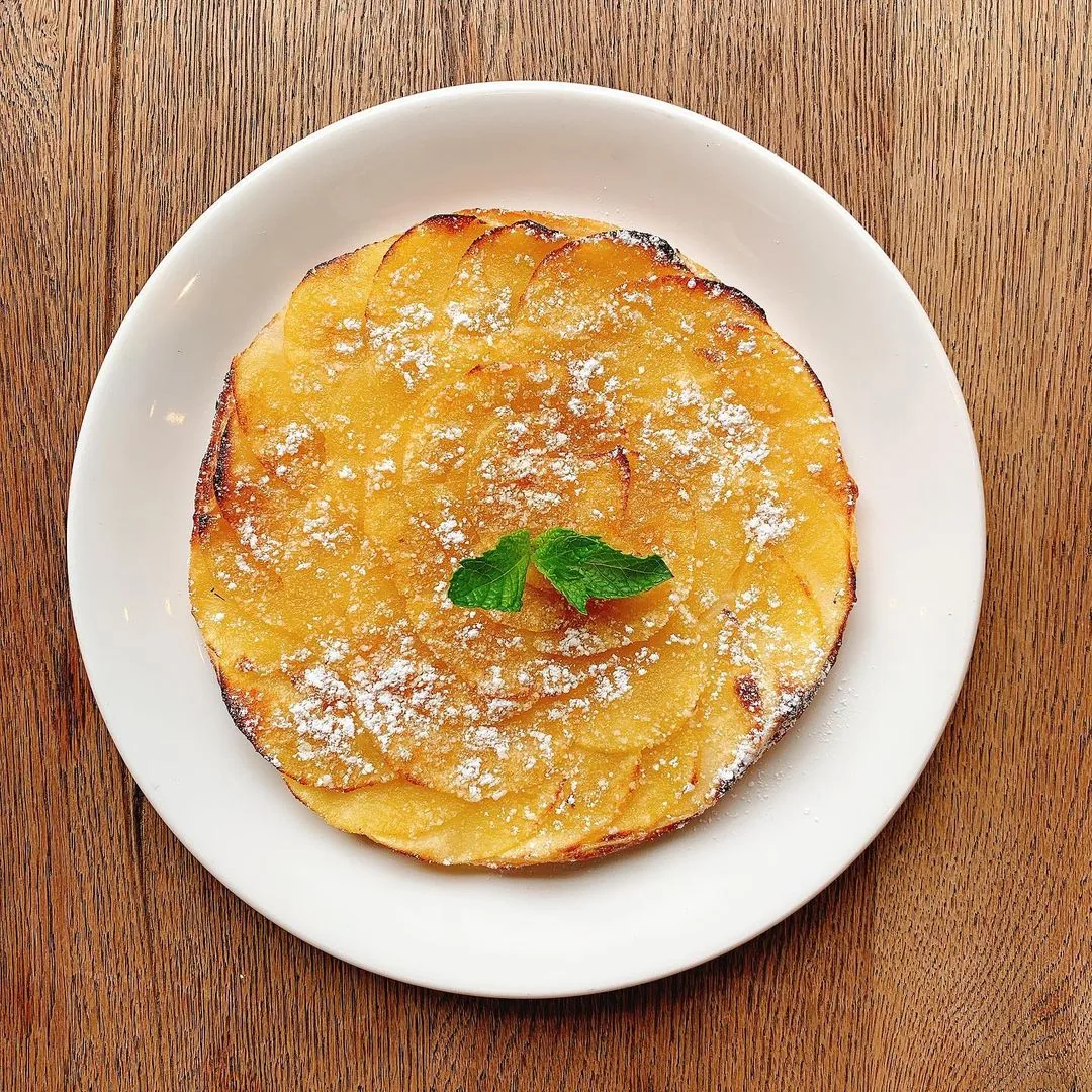 A thin apple tart with overlapping caramelised apple slices, dusted with icing sugar and topped with a sprig of mint, on a white plate