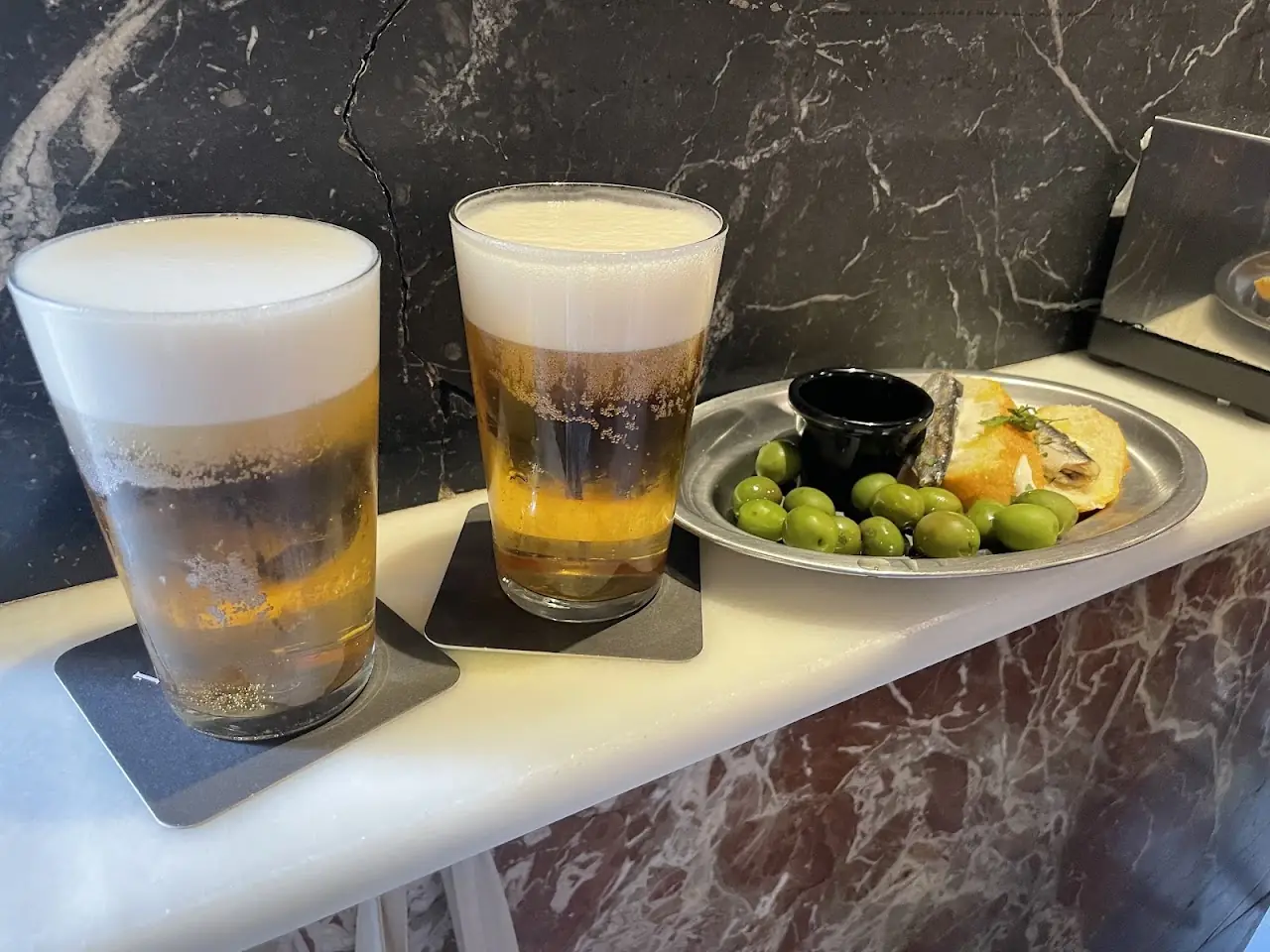 Two frothy glasses of beer next to a plate of olives and a small sandwich on the bar counter.