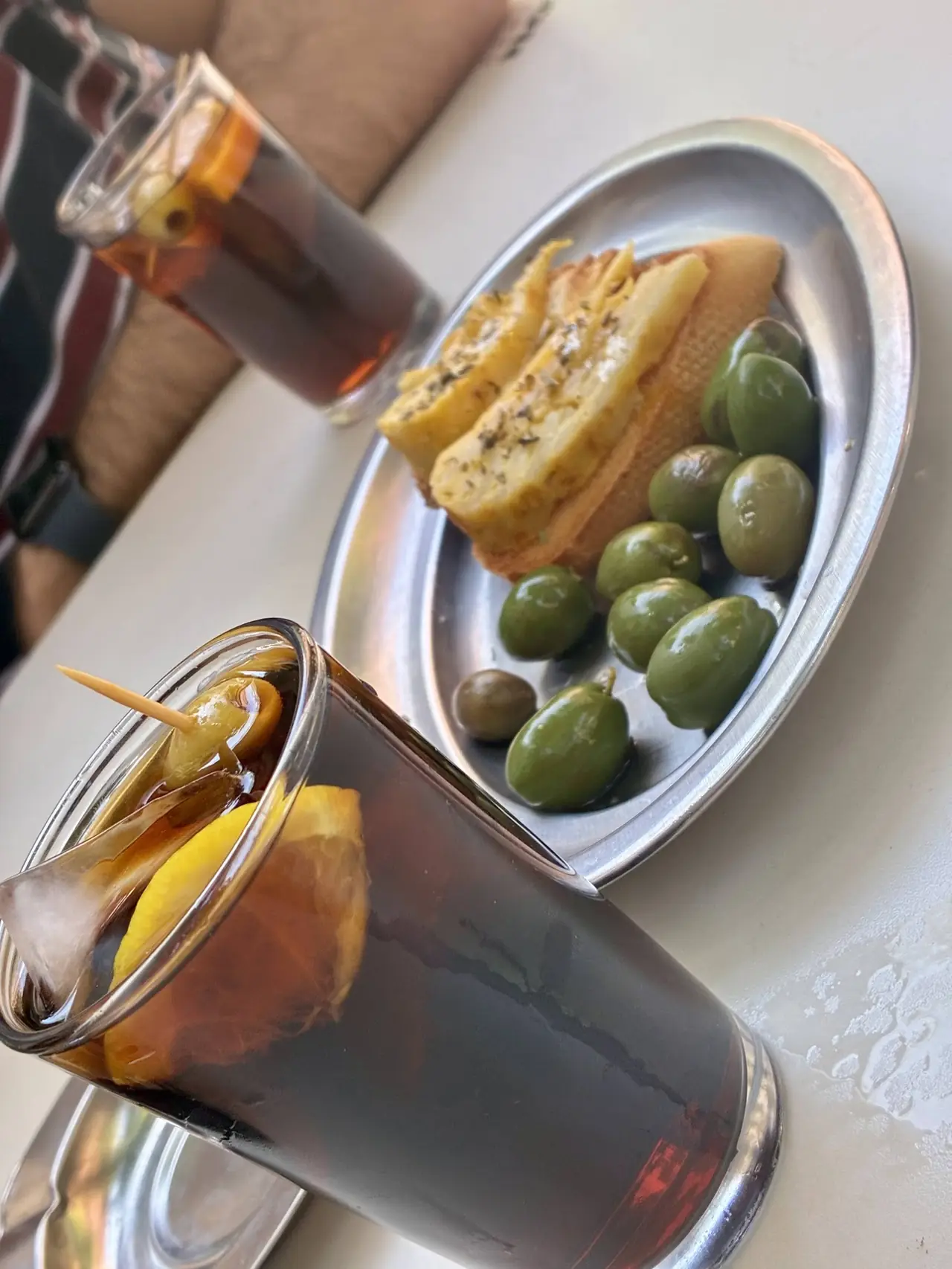 Two glasses of vermut with orange slice and olive garnish beside a plate of green olives and toasted bread