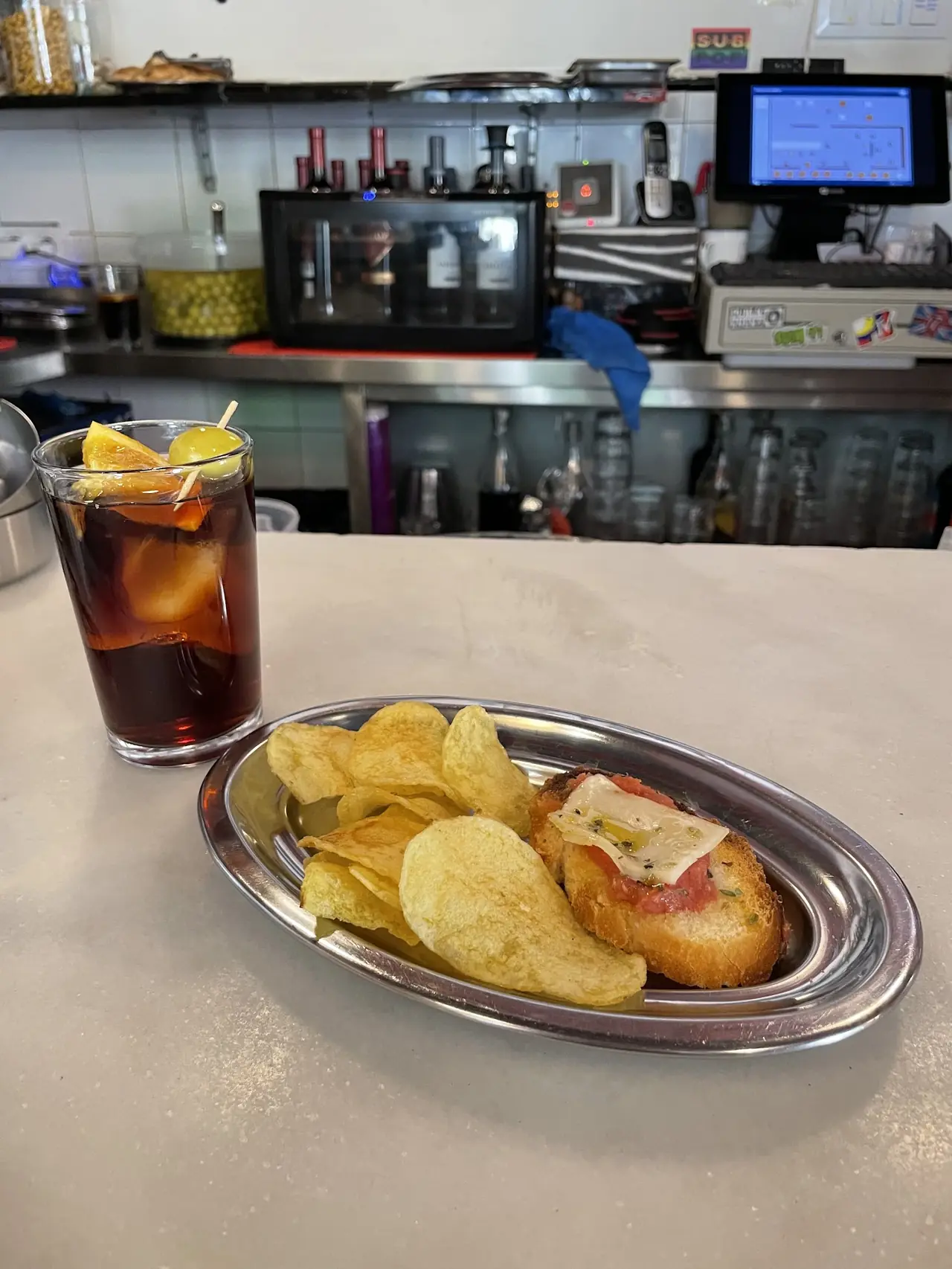 Glass of vermut with olive and orange garnish beside a plate of potato chips and a tostada with tomato and melted cheese