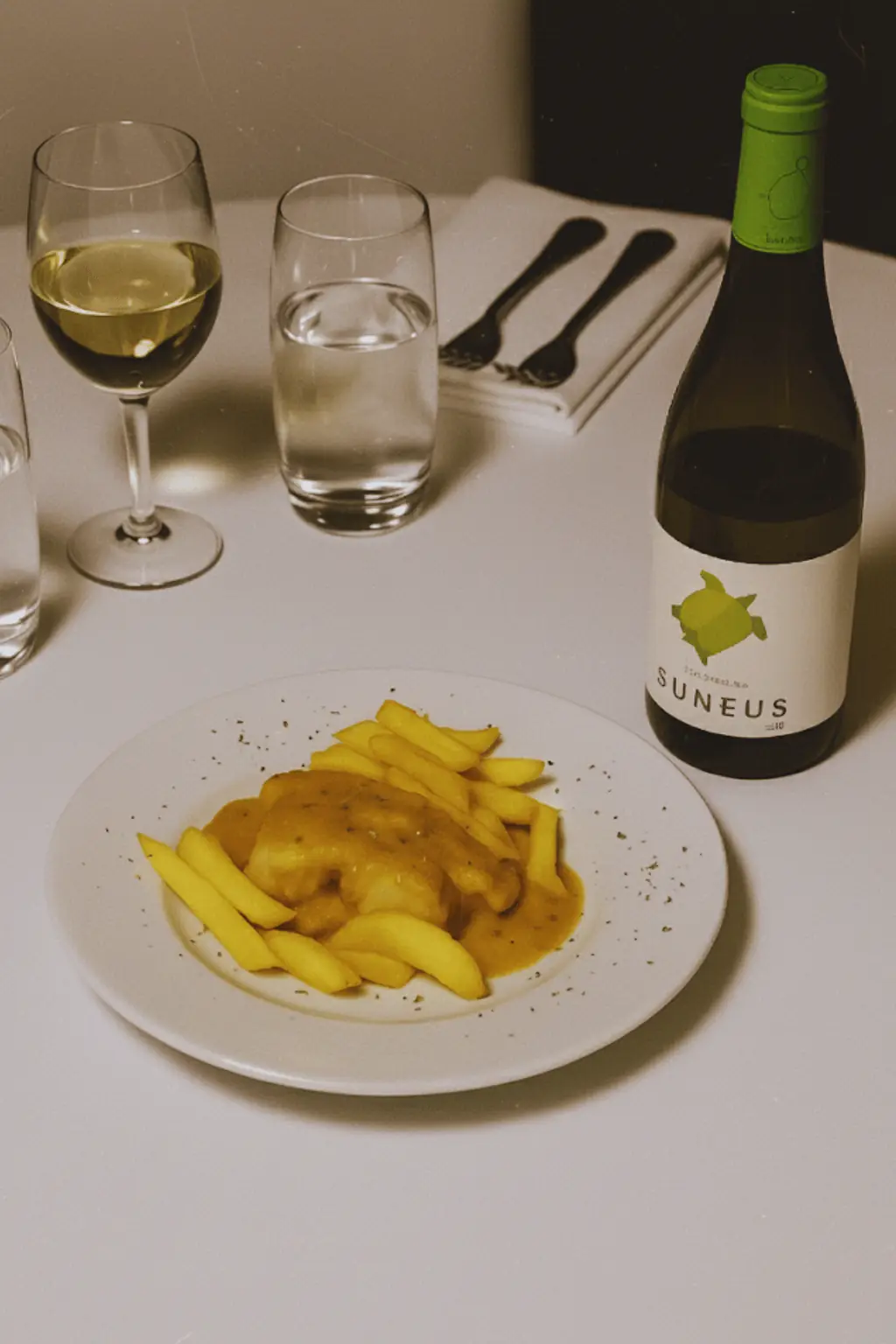 Battered fish with fried potato wedges in a savory sauce on a white plate, served with a glass of white wine, La Yaya Amelia