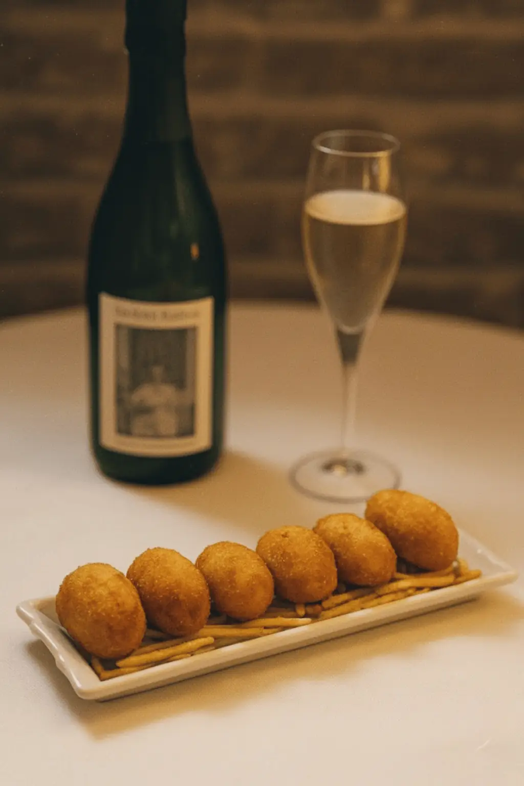 Row of breaded croquettes on a plate with matchstick potatoes, alongside a glass of sparkling wine, La Yaya Amelia