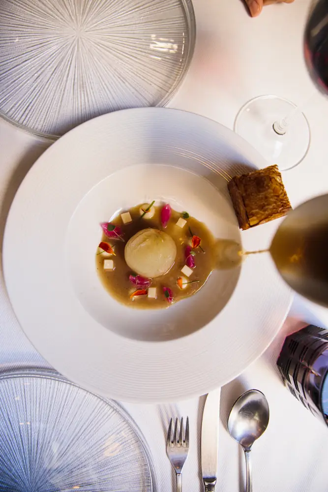 Fine-dining plate of a poached onion in brown broth being poured, garnished with tofu cubes and flower petals, with a crisp pastry square