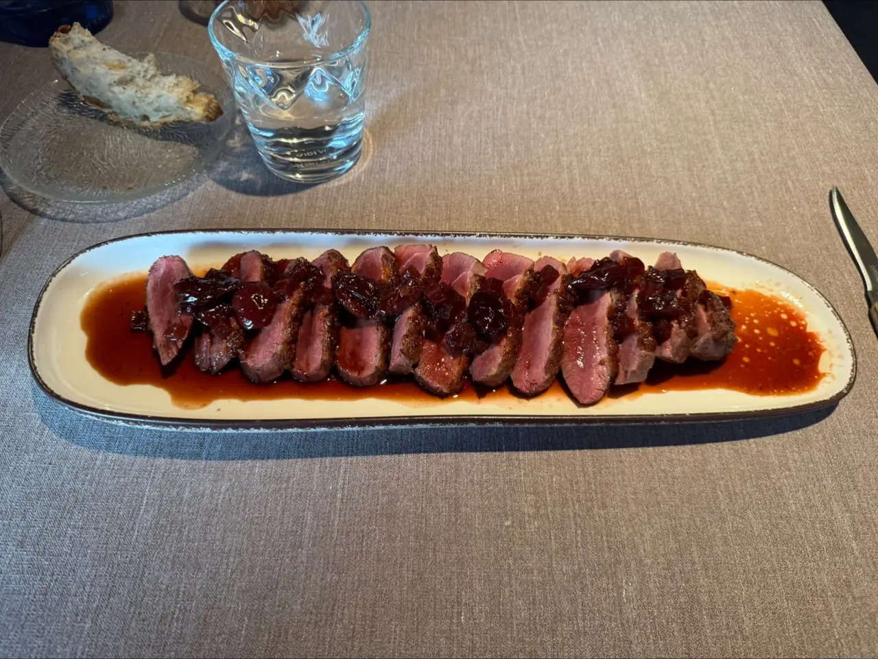 Long plate of sliced pink-cooked meat with a dark reduction sauce and dark fruit garnish, table setting in background