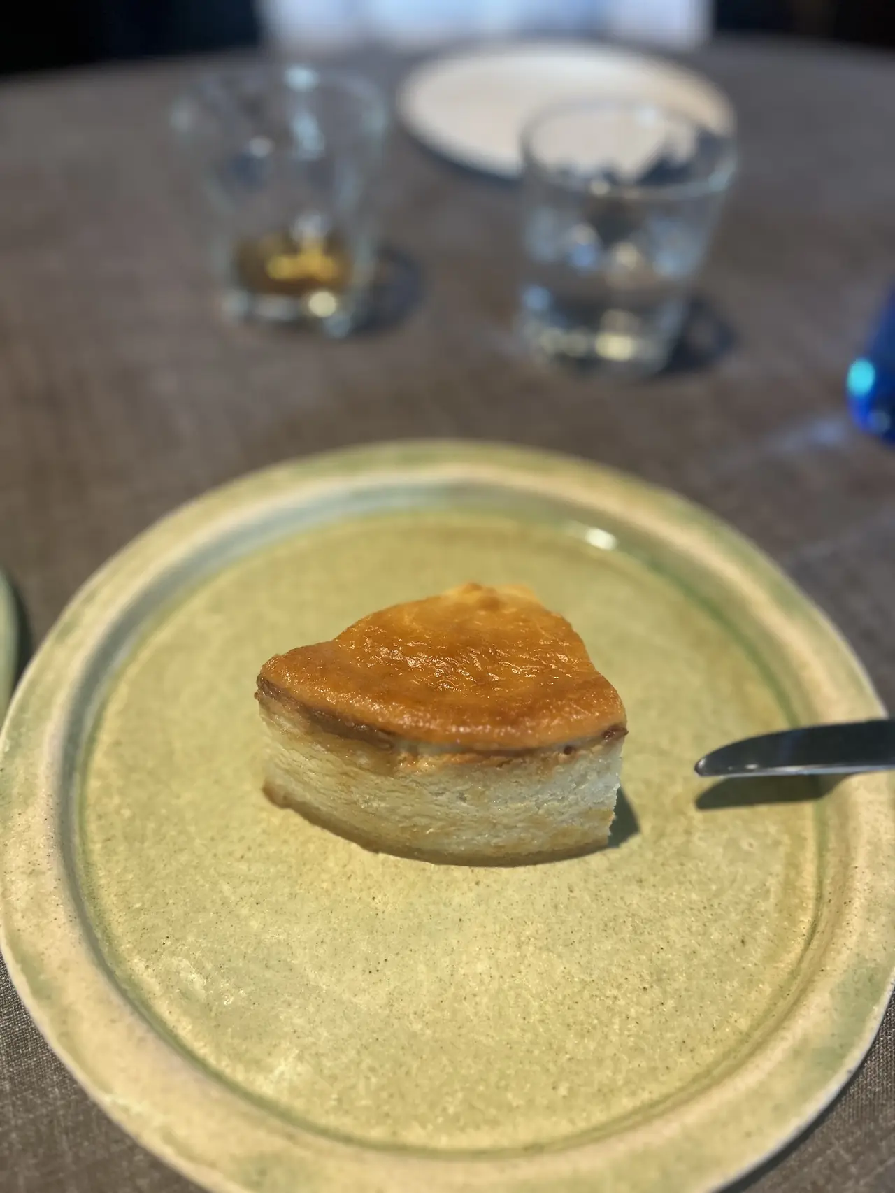 Slice of caramelized cheesecake on a plate with drinks blurred in the background