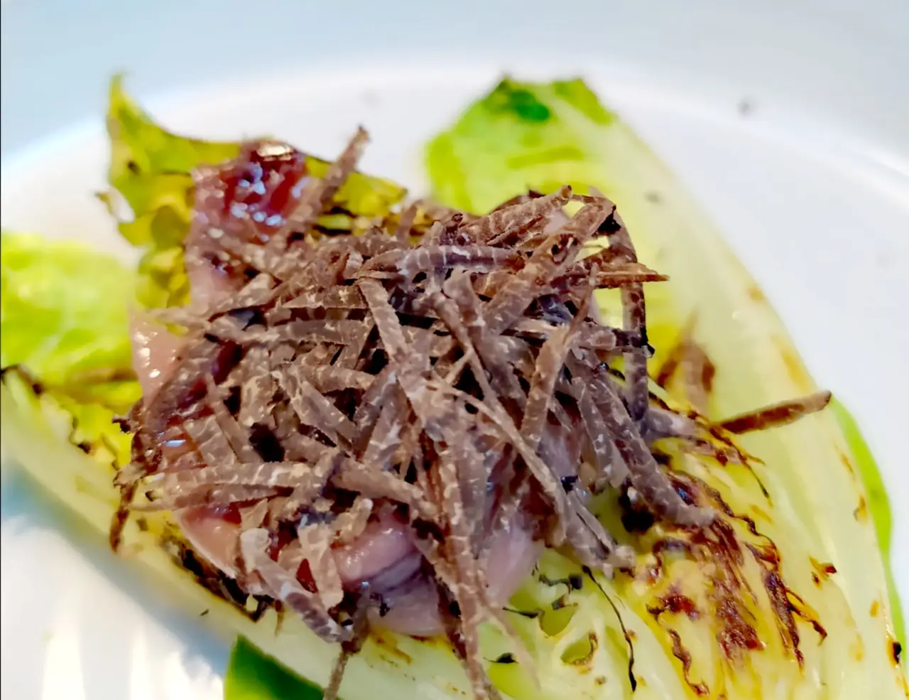 Close-up of grilled gem lettuce topped with shaved dried tuna and a piece of seared meat