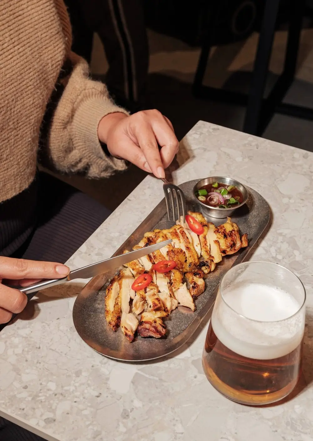 Sliced grilled chicken on a metal tray with sliced chilies and a dipping sauce, next to a glass of beer, Lamai Thai Tapas