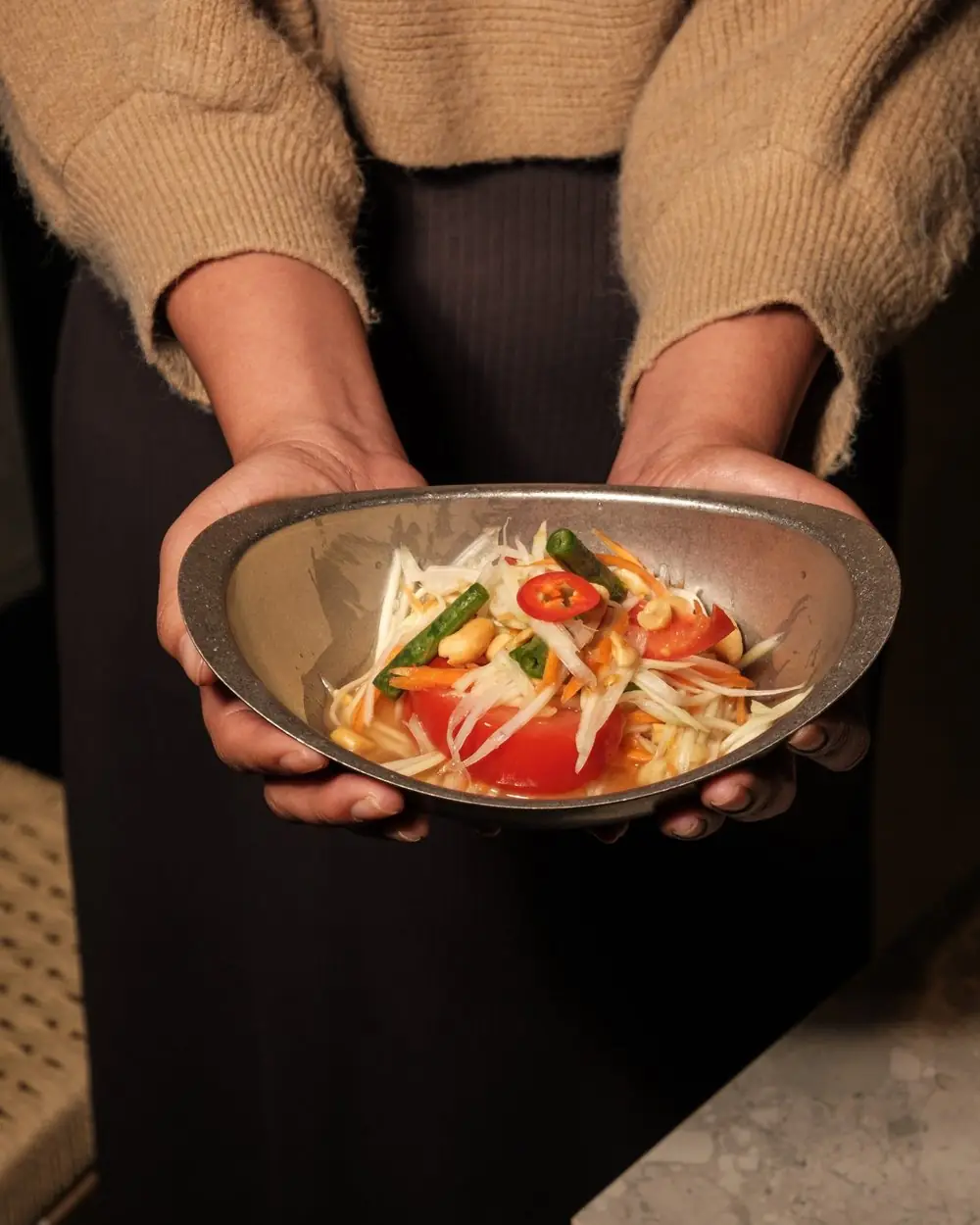 Green papaya salad with sliced tomato, peanuts, and chili in a metal bowl held in two hands, Lamai Thai Tapas