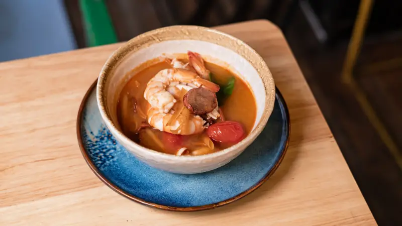 Shrimp soup with cherry tomatoes and herbs in a white bowl on a blue plate, at Lamun Cuina Thai