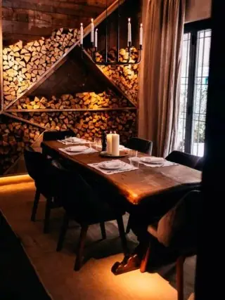 Long wooden table set for six in front of a wall of stacked firewood, lit by candles.