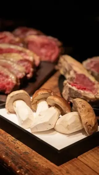Sliced fresh porcini on a white tray with racks of raw marbled beef behind on a wooden counter.