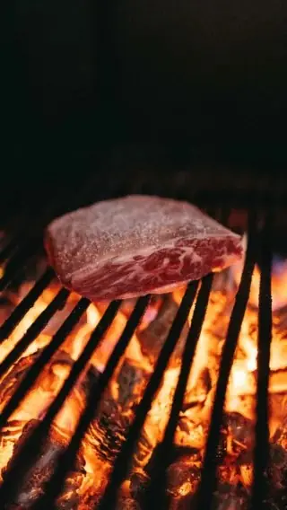 Thick marbled steak searing over a charcoal grill above glowing embers.