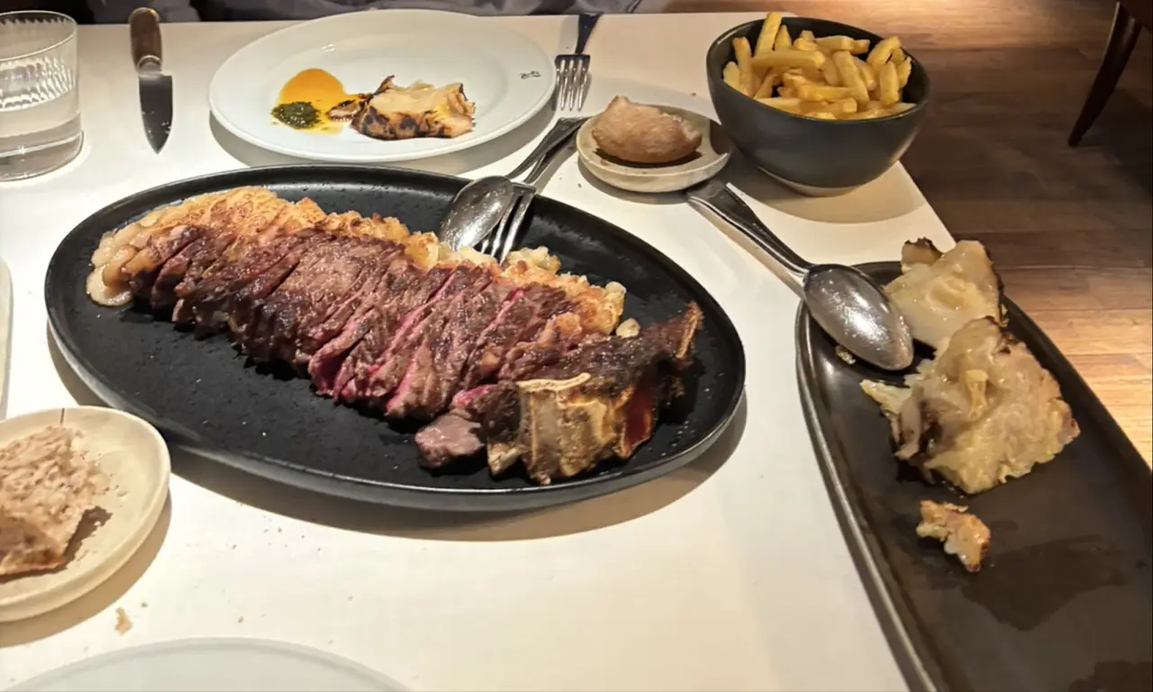 Sliced grilled steak on a black plate with a side of fries and other dishes on the table