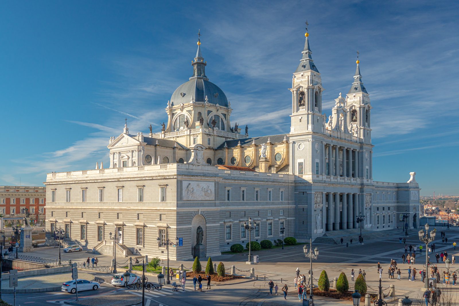 Almudena Cathedral's gray-and-white facade, dome, and twin bell towers rising over the plaza on a sunny day