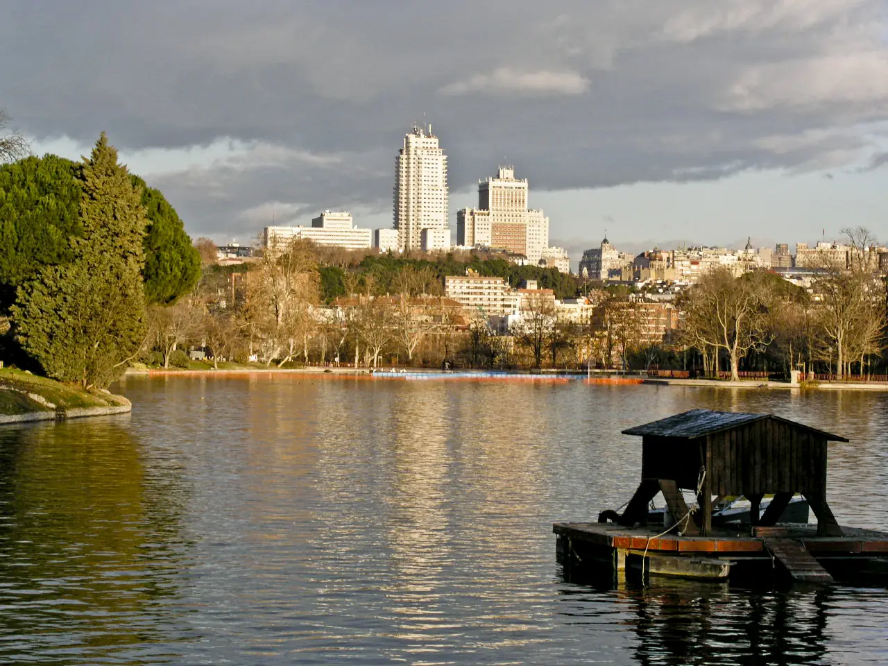 The lake at Casa de Campo with a wooden boathouse in the foreground and the Torre de Madrid and Edificio España skyline across the water